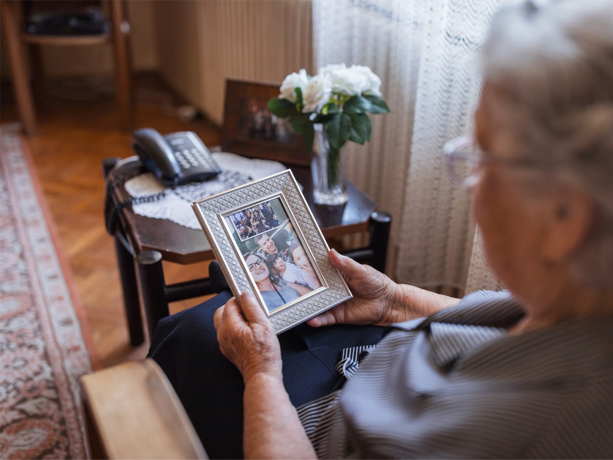 Older woman sitting in a cozy room in memory care, holding and looking at a framed family photo on her lap.