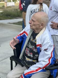 Bruce Beals, a 92-year-old Air Force veteran, sits in his wheelchair wearing a Honda jacket and motorcycle-themed shirt, smiling as he visits with others.